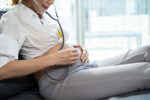 Asian beautiful pregnant woman sit on sofa and listen baby's heartbeat. Attractive young pregnancy mother feeling happy and excited while using stethoscope listen to newborn child vital signs in house