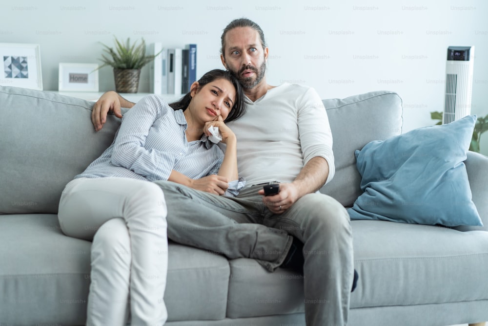 Caucasian loving couple watch movie together in living room at home. Beautiful happy new marriage man and woman sit on sofa use remote control TV and look to drama and sad show on television in house.