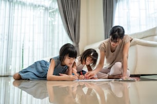 Asian Little siblings girl draw and color picture with mother on floor. Beautiful loving parent, mom spend time with small cute kid daughters enjoy activity at house. Parenting relationship concept.