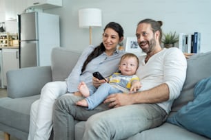Caucasian couple watching movie with baby kid in living room at home. Beautiful happy family sitting on sofa use remote control TV show and have fun laughing look to television while holding young son