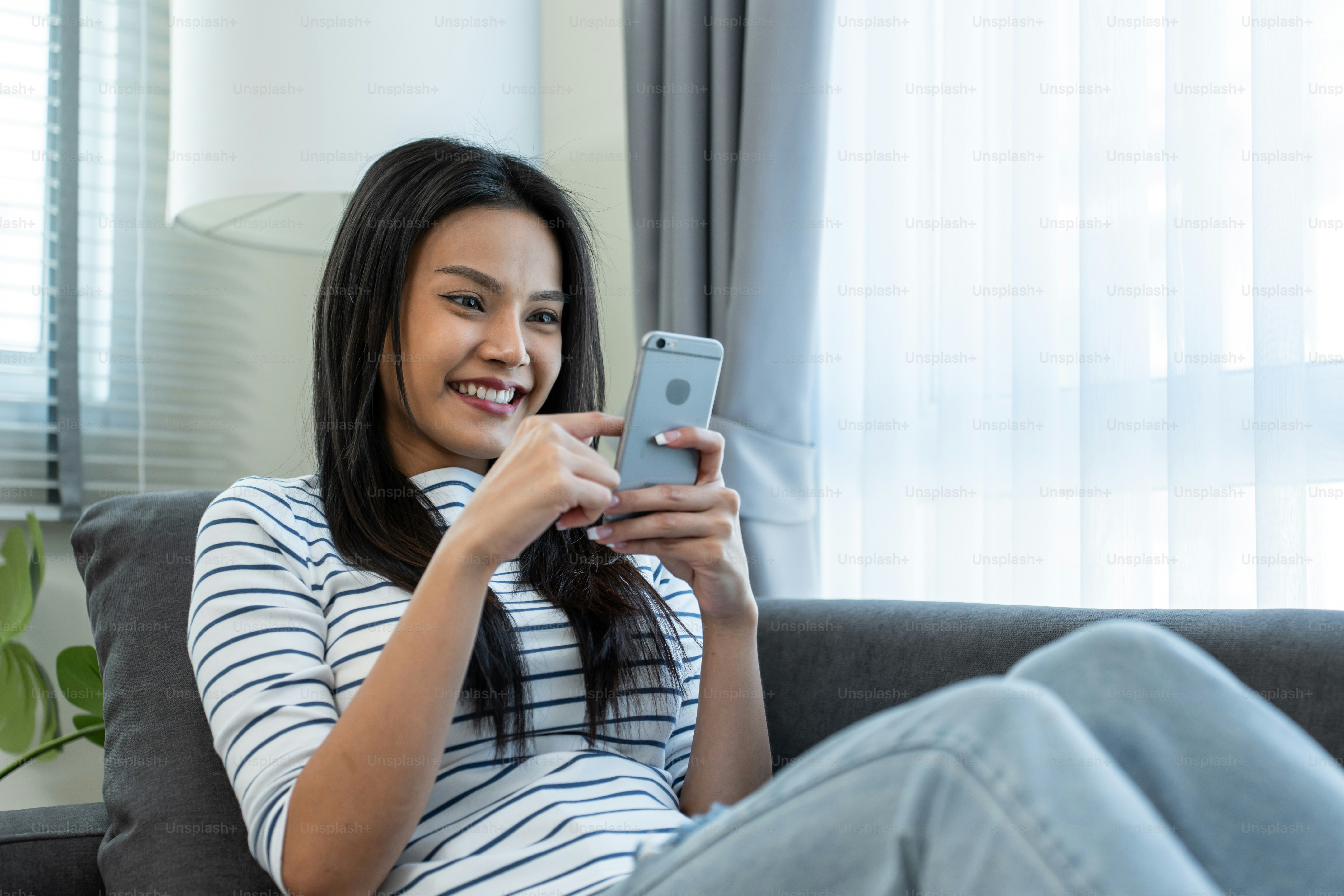 Belle femme asiatique s’assoit sur le canapé et discute sur le téléphone portable dans la maison. Heureuse jeune fille attrayante passer du temps libre à la maison, se sentir détendue et profiter de communiquer et discuter sur smartphone dans le salon.