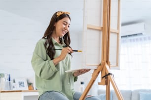 Asian young talented woman artist coloring on painting board in house. Attractive beautiful female draw art picture, creating artwork with watercolor paint and brush enjoy creativity activity at home.