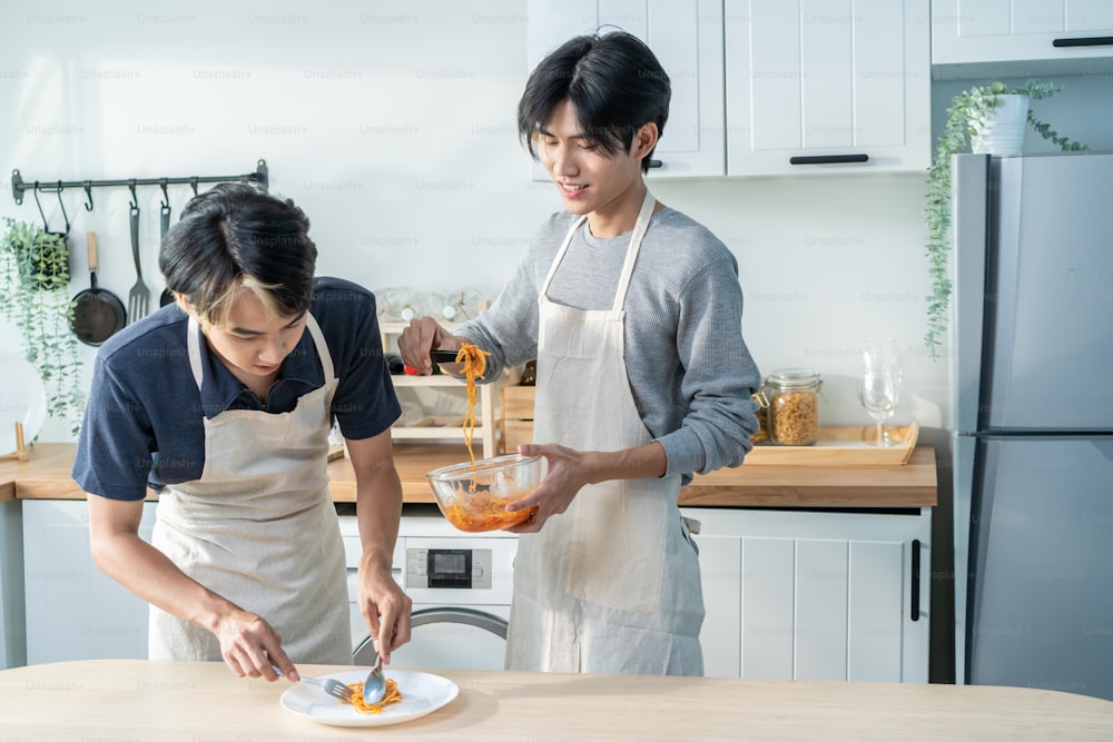 La giovane famiglia gay maschio LGBTQ asiatica si diverte a cucinare cibi in cucina a casa. Attraente bella coppia romantica dell'uomo indossa il grembiule sentendosi felice e allegro per trascorrere il tempo facendo gli spaghetti insieme in casa