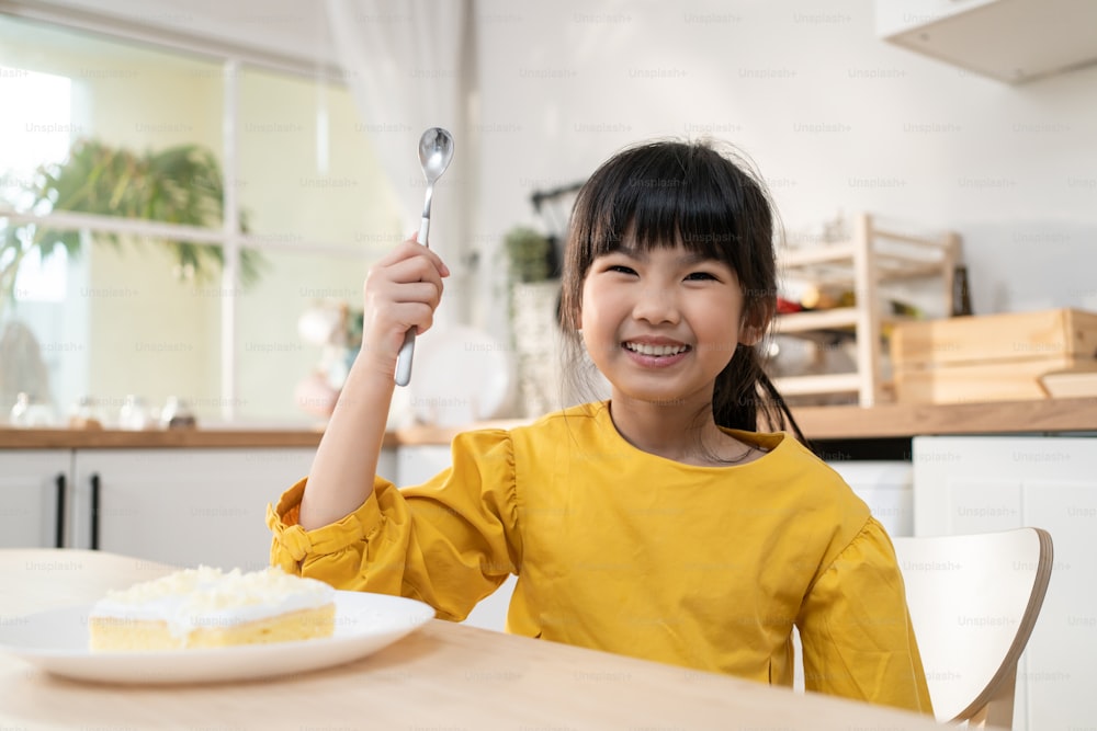 Portrait of Asian happy young kid girl eating cake in kitchen at home. Adorable little child sit on table feeling happy and enjoy sweet food after cooking foods or baking kneads yeast dough in house.