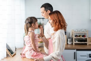 Asian attractive parents feed cupcake to cute baby daughter in kitchen. Happy family, loving couple baking and serve tasty sweet foods to little cute kid girl while sit on counter for lunch in house.