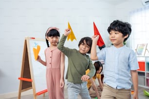 Grupo de niños pequeños de raza mixta que juegan al avión en el aula de la escuela. Los niños, niñas y niños, estudiantes que pasan tiempo jugando cohetes de papel durante el recreo, se divierten juntos en el aula de la escuela.