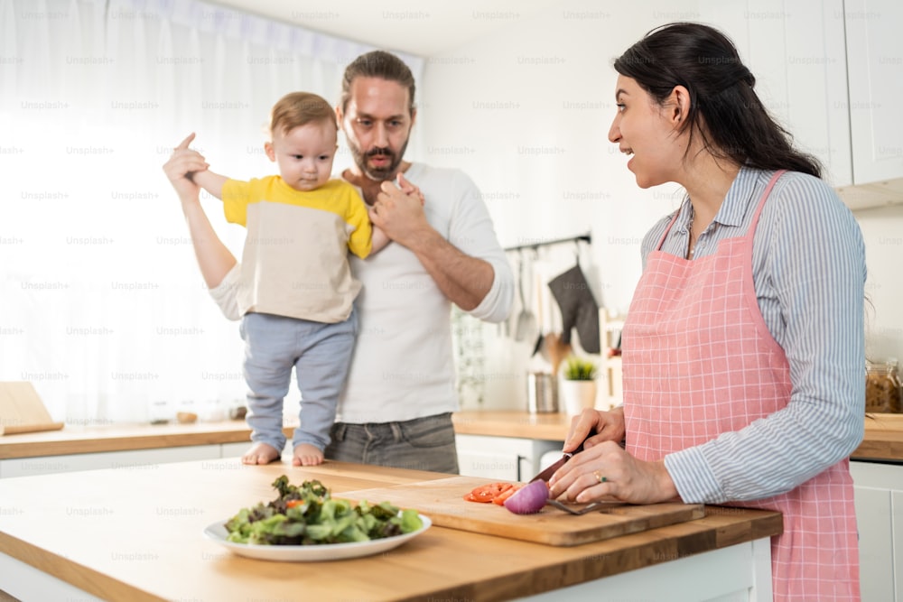 Caucasian beautiful parents cook food with baby boy toddler in kitchen. Happy family, Attractive young mother making healthy salad for lunch while husband father holding little kid son infant in house