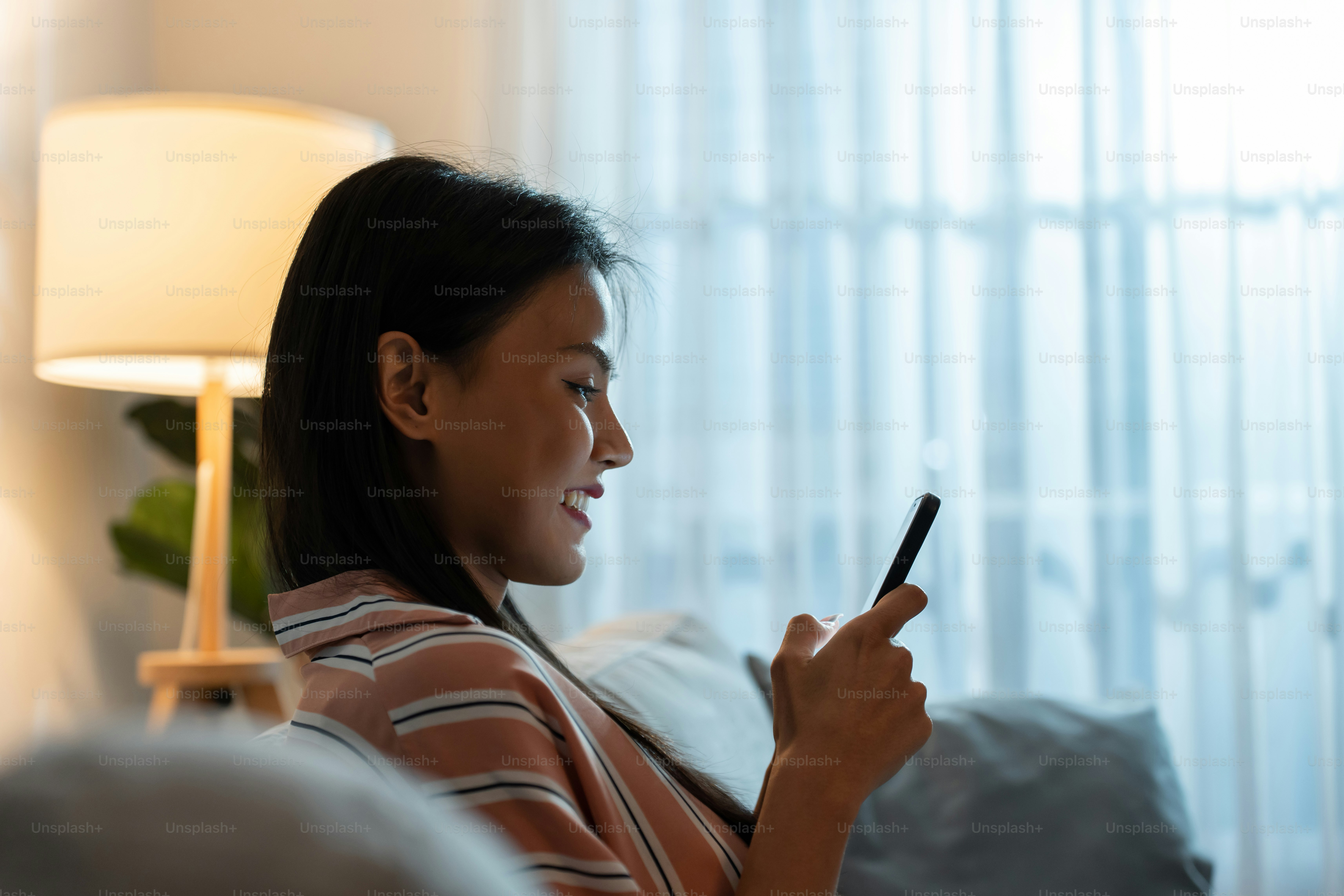Belle femme asiatique s’assoit sur le canapé et discute sur le téléphone portable la nuit. Jolie jeune fille heureuse passer du temps libre à la maison, se sentir détendante et profiter de communiquer et discuter sur smartphone dans le salon.