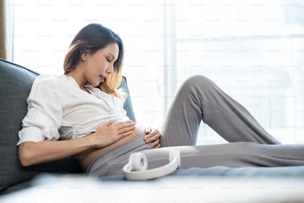 Jovem asiática bela mulher grávida aplicando fones de ouvido em sua barriga. Atraente grávida menina mãe sentar-se no sofá dentro de casa na sala de estar e tocar música para o bebê recém-nascido na barriga para ouvir.