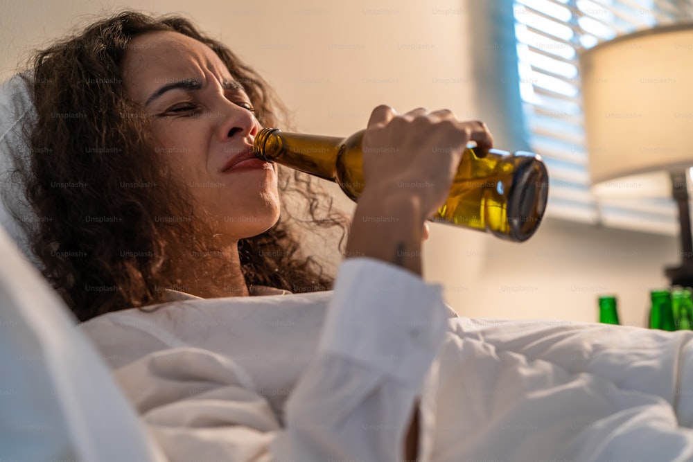 Una mujer latina borracha sostiene una botella de cerveza y llora y siente resaca en la cama. Joven hermosa mujer de depresión que miente y llora llorando en el dormitorio, siente el corazón roto y celebra la vida con alcohol por la noche
