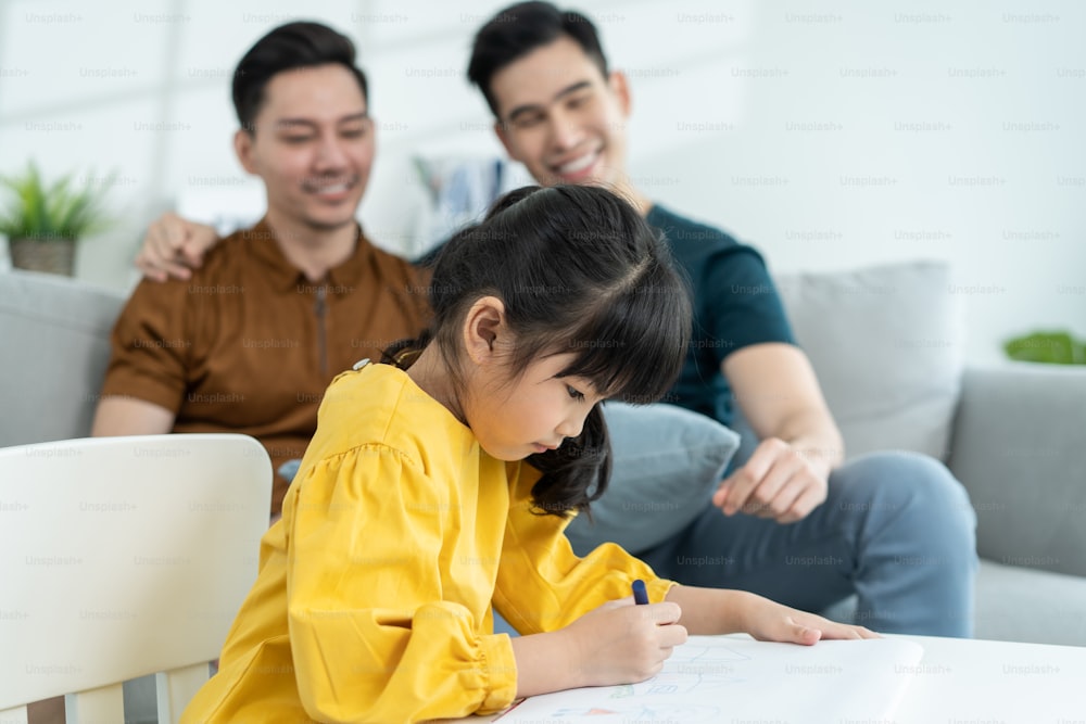 Asian attractive LGBTQ gay family teach young girl kid draw picture. Handsome male couple look at little adorable child daughter play and coloring book in living room, enjoy parenting activity at home