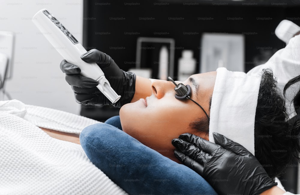 Hände eines Profis, der einen Laser zur Hautpflege eines jungen lateinamerikanischen Mannes verwendet.