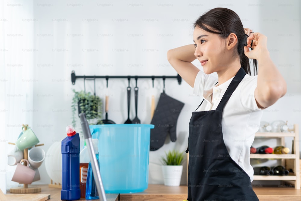 Employée de service de nettoyage asiatique attachant les cheveux avant de faire le ménage. Belle femme de ménage jeune fille femme de ménage se sentir heureuse et se préparer à nettoyer la table de cuisine pour le ménage ou les tâches ménagères travaillant à la maison