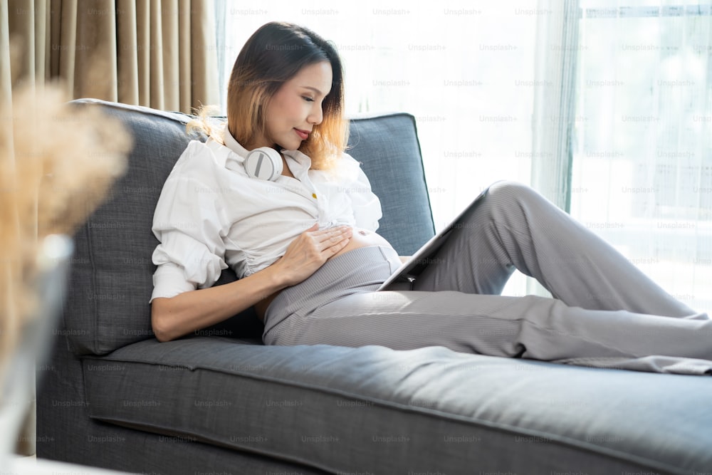 Joven asiática hermosa mujer embarazada sosteniendo y acariciando su barriga. Atractiva madre embarazada embarazada sentada en el sofá de la sala de estar de su casa y mirando el vientre con felicidad y amor.