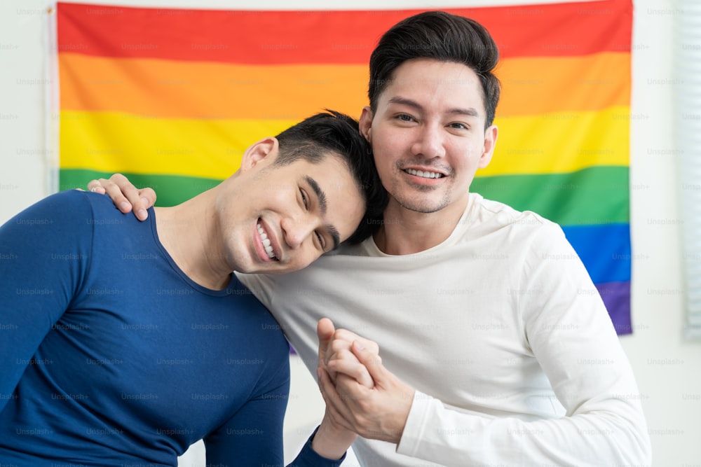 Ritratto di bella famiglia gay uomo asiatico che tiene la bandiera e il sorriso LGBT. Attraente coppia lgbt maschile romantica si siede sul letto in camera da letto al mattino e guarda la telecamera con orgoglio gay e sfondo arcobaleno