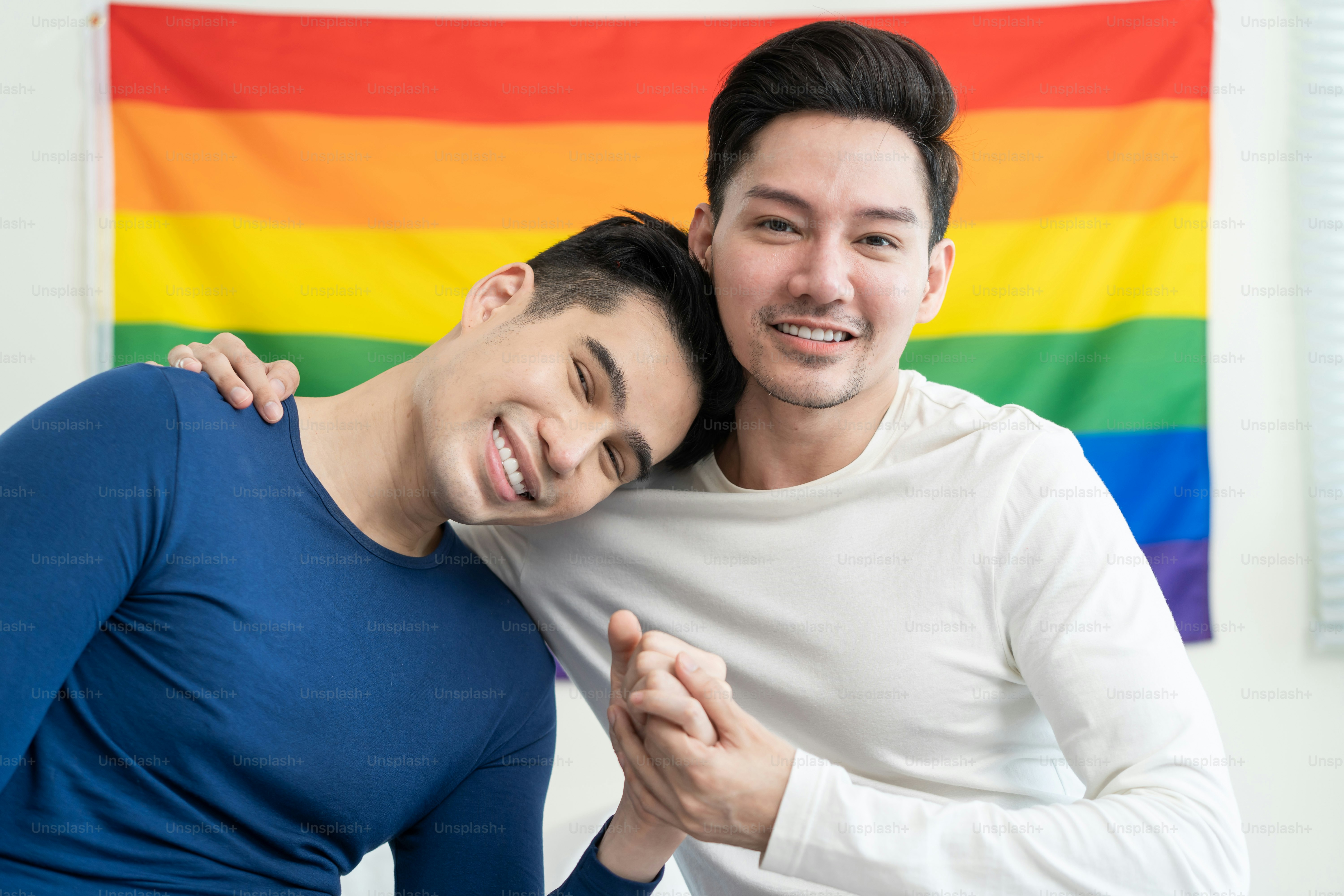 Two men in long-sleeved shirts holding hands in front of a rainbow flag