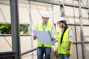 Asian Colleagues worker Specialists team wearing protective safety helmet look at blueprint on Construction Site. Civil Engineer Male and architect discuss about Real Estate Building Project work plan