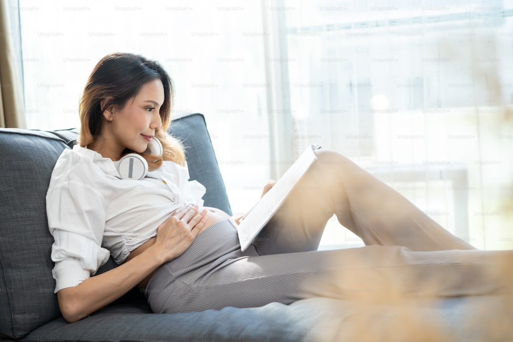 Giovane donna incinta asiatica che applica le cuffie sulla pancia. La madre attraente della ragazza in attesa di gravidanza si siede sul divano all'interno del soggiorno e suona la musica per il neonato nella pancia da ascoltare.
