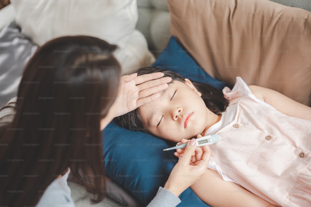 Hija enferma de una niña asiática durmiendo en el sofá con fiebre mientras la madre controla la temperatura en su frente