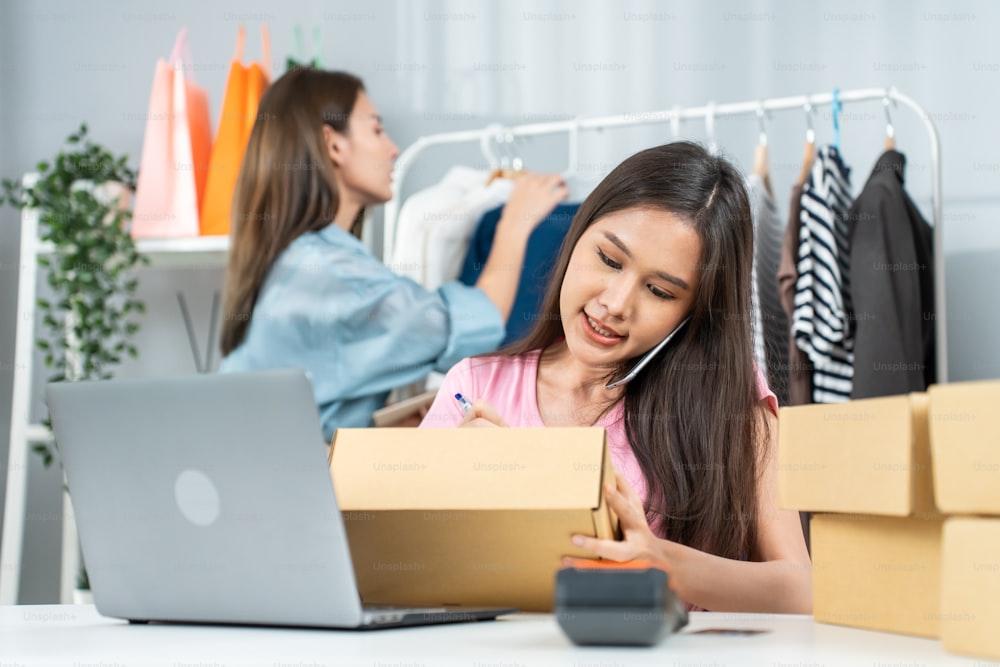 Asian woman friends couple packing clothes order into box for customer. Young attractive business people work to preparing parcel boxes check ecommerce shipping online retail to sell at office store.