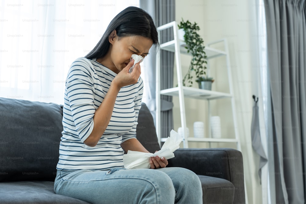 Asian beautiful woman having running nose and sneezing in living room. Attractive young female sitting on sofa feeling bad and suffer from allergy then put tissue cover her nose while sneez in house.