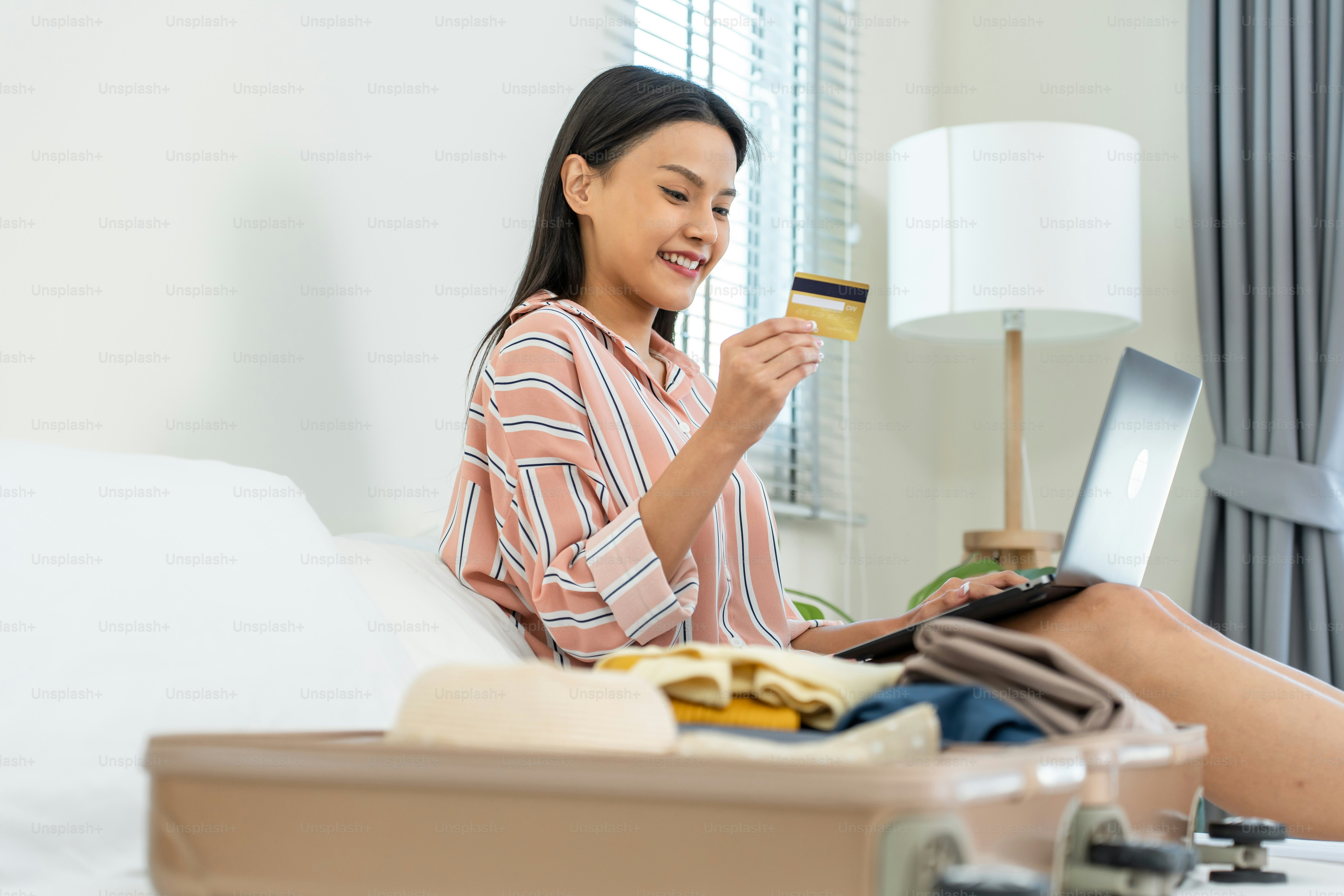 Asian young woman use credit card to pay for booking hotel in bedroom. Attractive female tourist traveler feel happy and relax while preparing luggage on bed, ready to travel on holiday vacation trip.