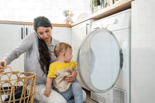 Caucasian busy mother doing housework with baby boy toddler in kitchen. Beautiful mom use phone call for work and put clothes to washing machine with her son play around in house. Family-housekeeping.