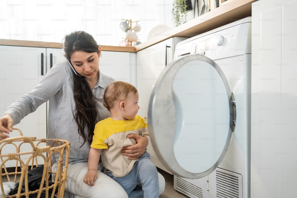 Caucasian busy mother doing housework with baby boy toddler in kitchen. Beautiful mom use phone call for work and put clothes to washing machine with her son play around in house. Family-housekeeping.