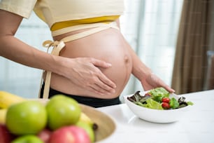 Primer plano de una mujer asiática embarazada de pie y sosteniendo un cuenco de mezcla. Atractiva y hermosa mujer joven embarazada de pie y tocando al bebé en el útero, elija comida saludable o comida vegetariana en la cocina.