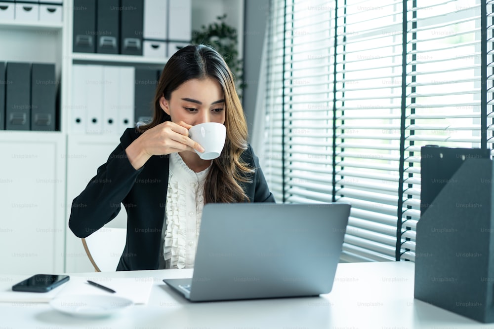 Asian beautiful business woman smile drink cup of coffee work in office. Attractive professional female employee worker in formal wear sit on table at workplace, use laptop computer enjoy in corporate