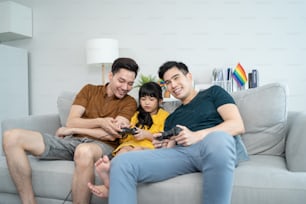 Asian gay couple play video game with young daughter in living room. Little adorable girl child sit on sofa with attractive romantic young male LGBTQ family enjoy using joystick and play game together
