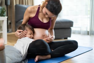 Asian young lovely daughter hugging beautiful pregnant mother tummy. Attractive expecting pregnancy girl embrace little girl with happiness and love while doing yoga exercise in living room at home.