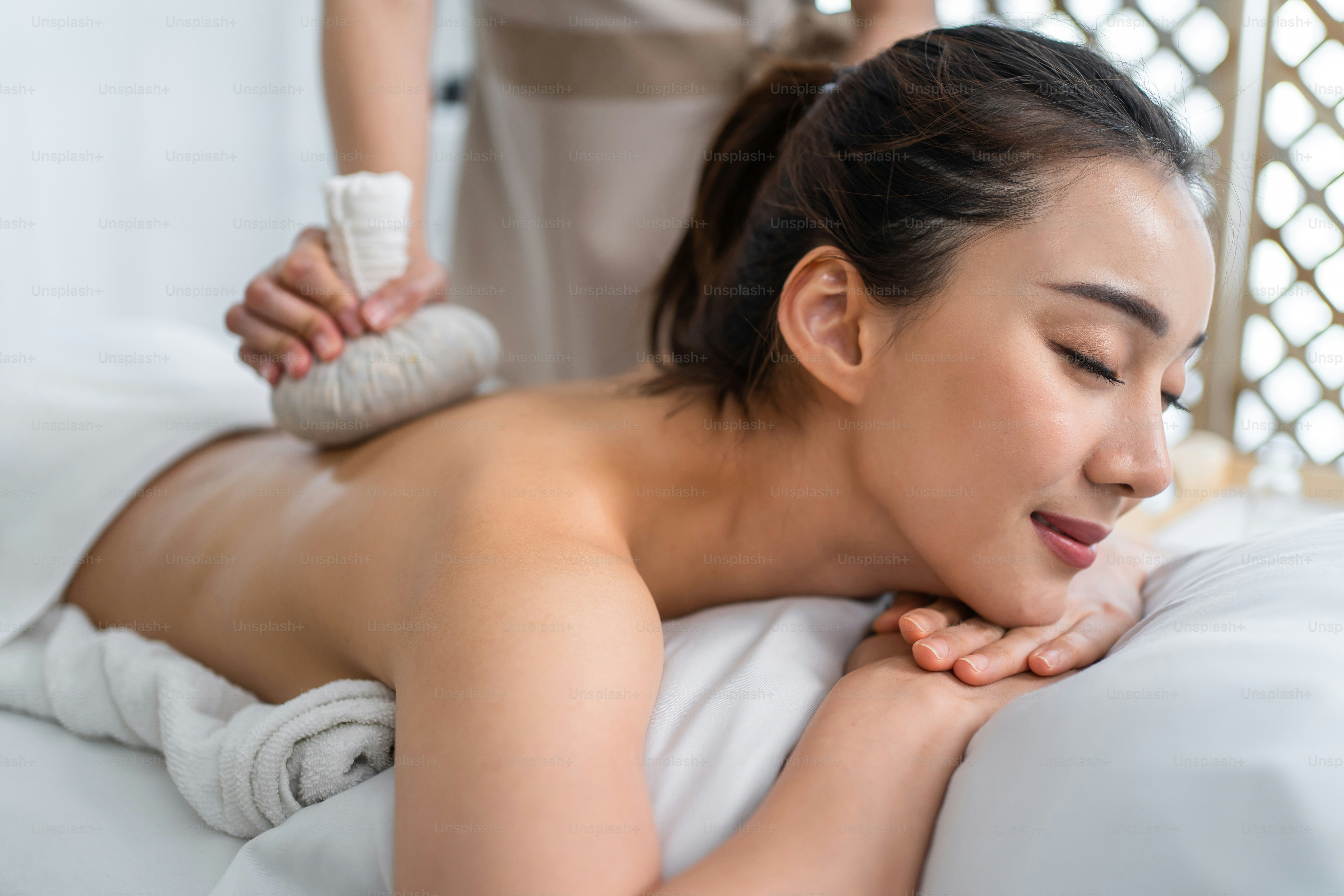 Wanita cantik Asia menikmati pijat kompres panas Thailand dengan tas herbal. Gadis menarik berbaring di atas meja pijat, merasa bahagia dan rileks selama fisioterapi punggung dari tukang pijat untuk perawatan tubuh di salon spa.