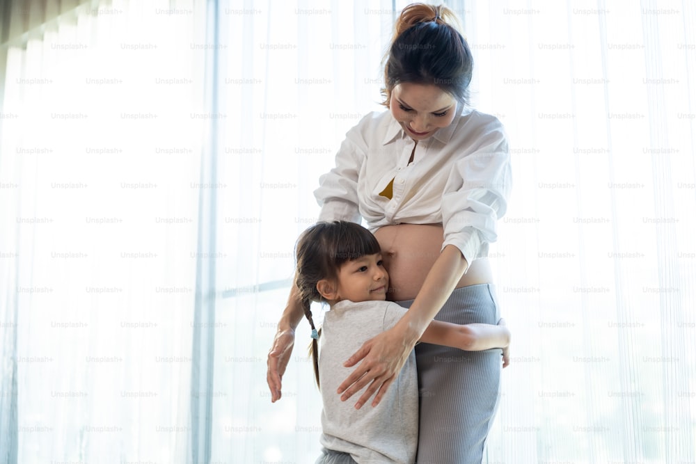 Asian young daughter hug and stroking beautiful pregnant mother tummy. Attractive expecting pregnancy girl embrace little kid girl with happiness and love while stand indoor near window in living room