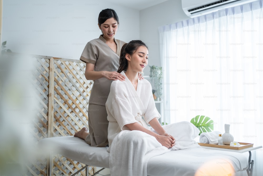 Joven asiática feliz que se siente relajada durante el masaje de hombros y espalda. Atractiva chica hermosa sentada en la mesa de masaje que obtiene servicio de fisioterapia de masajista para el cuidado del cuerpo en el salón de belleza del spa