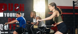 Group of young athlete people doing exercise together in fitness gym. Attractive active sportsman and women work out together by cycling on bike machine for health care and well being in stadium.