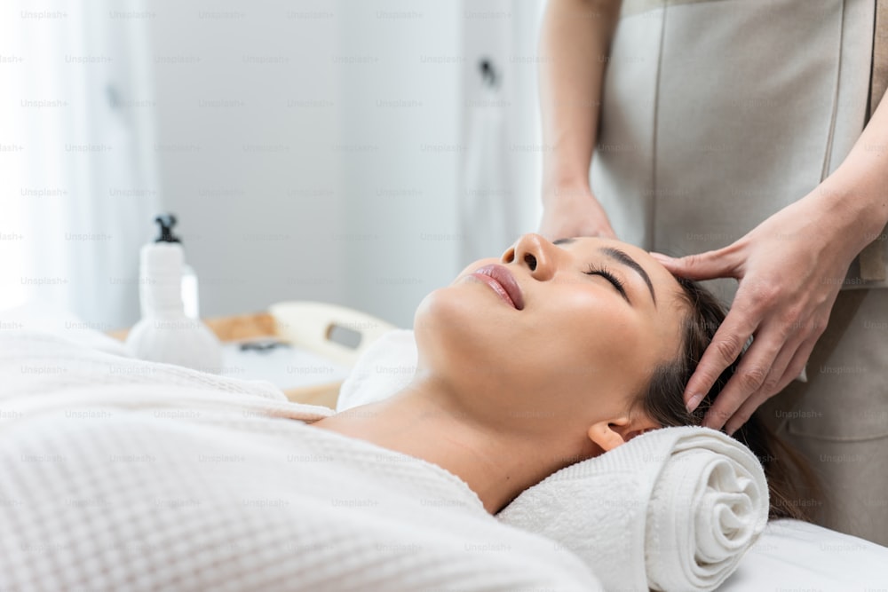 Mulher jovem asiática sentir-se feliz e relaxar durante a massagem facial e cabeça. Menina bonita atraente deitada na mesa de massagem, recebendo fisioterapia de massagista para cuidados com a pele e o corpo no salão de beleza do spa.