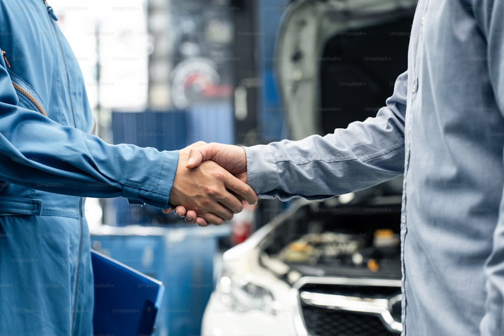 ガレージでクライアントと握手するアジアの自動車整備士の修理工。メカニックワークショップで働く車両サービスマネージャーは、顧客の車のエンジンをチェックし、メンテナンスした後、幸せと成功を感じます。