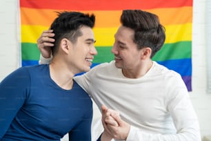 Portrait of Asian handsome man gay family holding LGBT flag and smile. Attractive romantic male lgbt couple sit on bed in bedroom in morning, look at each other with gay pride and rainbow background.