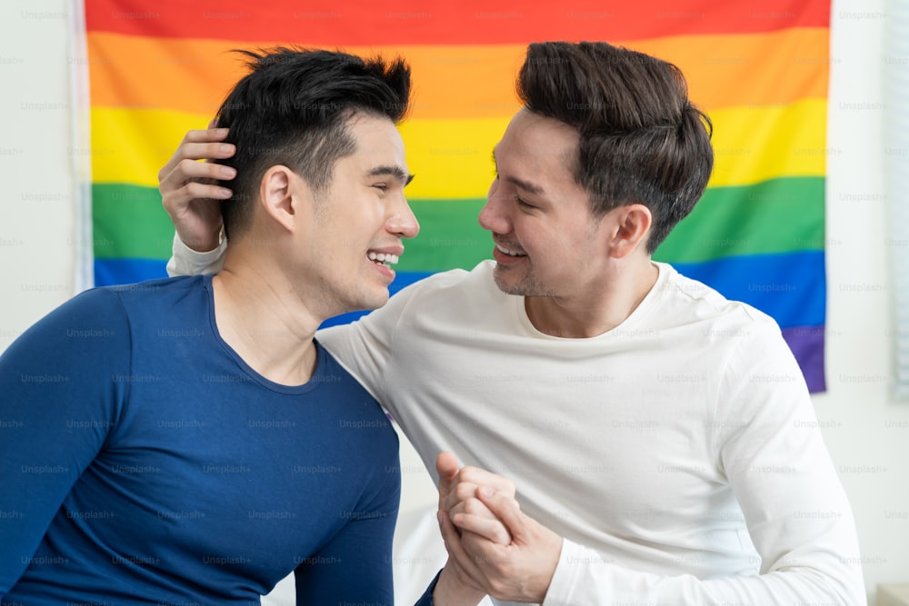 Ritratto di bella famiglia gay uomo asiatico che tiene la bandiera e il sorriso LGBT. Attraente coppia lgbt maschile romantica si siede sul letto in camera da letto al mattino, si guarda con orgoglio gay e sfondo arcobaleno.