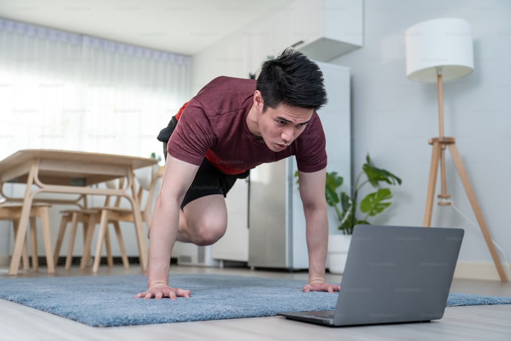 Asian handsome active young man doing exercise on floor in living room. Attractive male in sportswear spend leisure activity time to workout for health by follow online instruction on laptop at home.