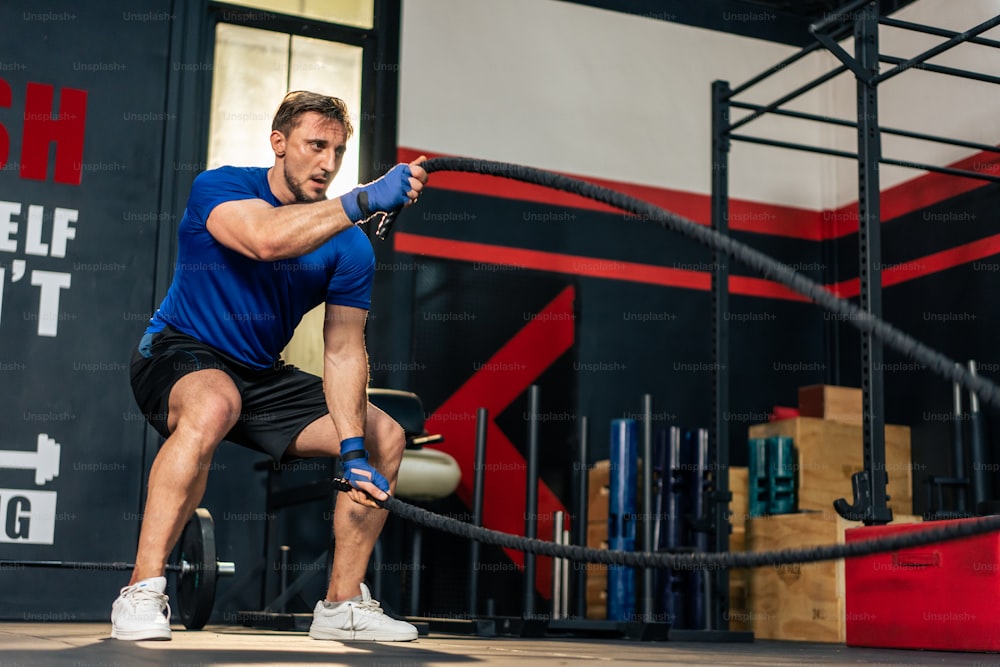 Caucasian active handsome sportsman working out in fitness gym club. Attractive athlete male exercise using battle rope to lose weight and maintain muscle for health care lifestyle in fitness stadium.