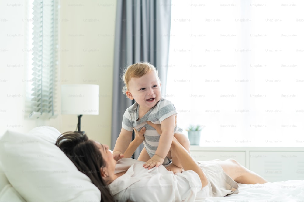 Caucasian loving mother playing with baby boy child on bed in bedroom. Happy family, attractive beautiful caring young mother lifting toddler son fly up enjoy doing airplane game in morning in house.