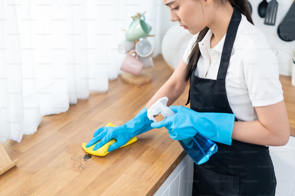 Asian young cleaning service woman worker clean kitchen table at home. Beautiful young girl housekeeper cleaner feel happy and wiping messy dirty cooking counter for housekeeping housework or chores.