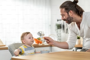Caucasian beautiful father take care of baby boy toddler in kitchen. Happy family, Attractive loving daddy cook and feeding healthy foods to little kid son infant while sit on chair for lunch in house