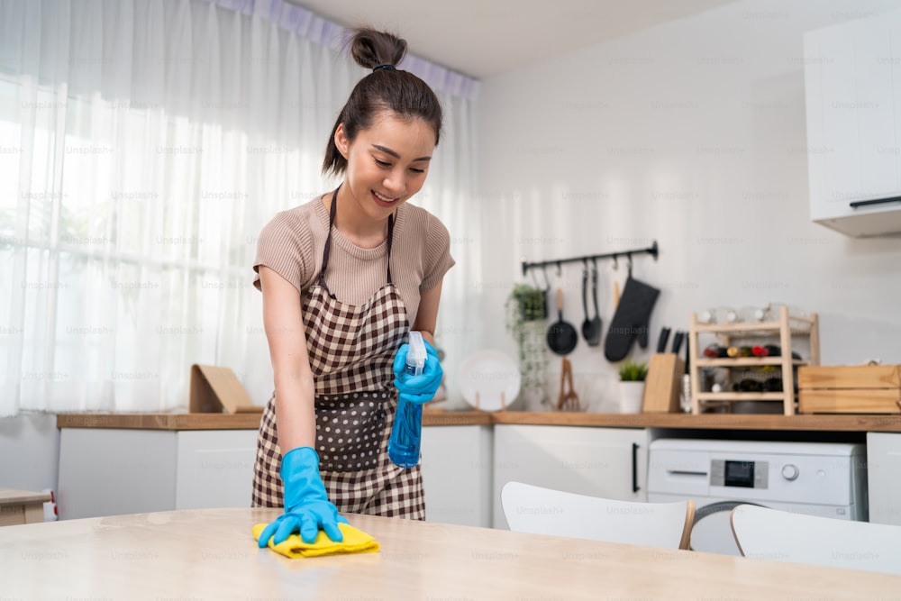 Joven asiática trabajadora del servicio de limpieza limpia la mesa de la cocina en casa. La hermosa joven ama de llaves limpiadora se siente feliz y limpia la encimera de cocina sucia y desordenada para las tareas domésticas o las tareas domésticas.