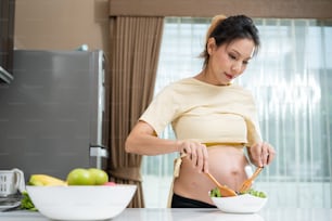 Asian beautiful pregnant woman cooking healthy food in kitchen at home. Attractive pregnancy girl stand indoors in kitchen feel happy and enjoy making green vegetable salad in bowl for health in house