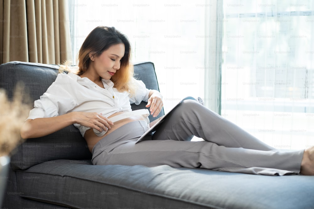 Giovane donna asiatica incinta seduta e con le cuffie sulla pancia. La gravidanza femminile attraente si siede sull'apertura del divano di comfort che suona la musica al bambino nel grembo materno con felicità e sorridente nel soggiorno di casa