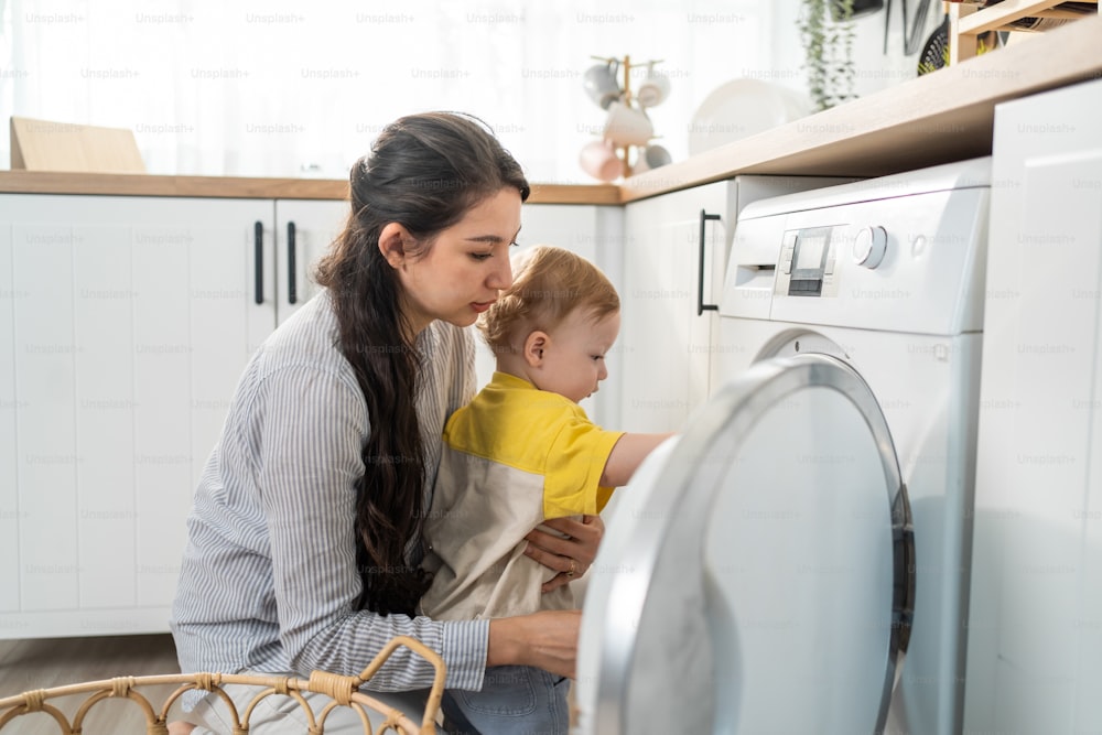 Caucasian young mom doing housework with baby boy toddler in kitchen. Beautiful mother doing laundry, put clothes to washing machine while take care of her son little kid in house. Family-housekeeping