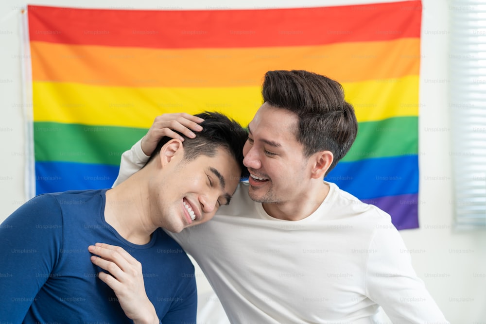 Portrait of Asian handsome man gay family holding LGBT flag and smile. Attractive romantic male lgbt couple sit on bed in bedroom in morning, look at each other with gay pride and rainbow background.
